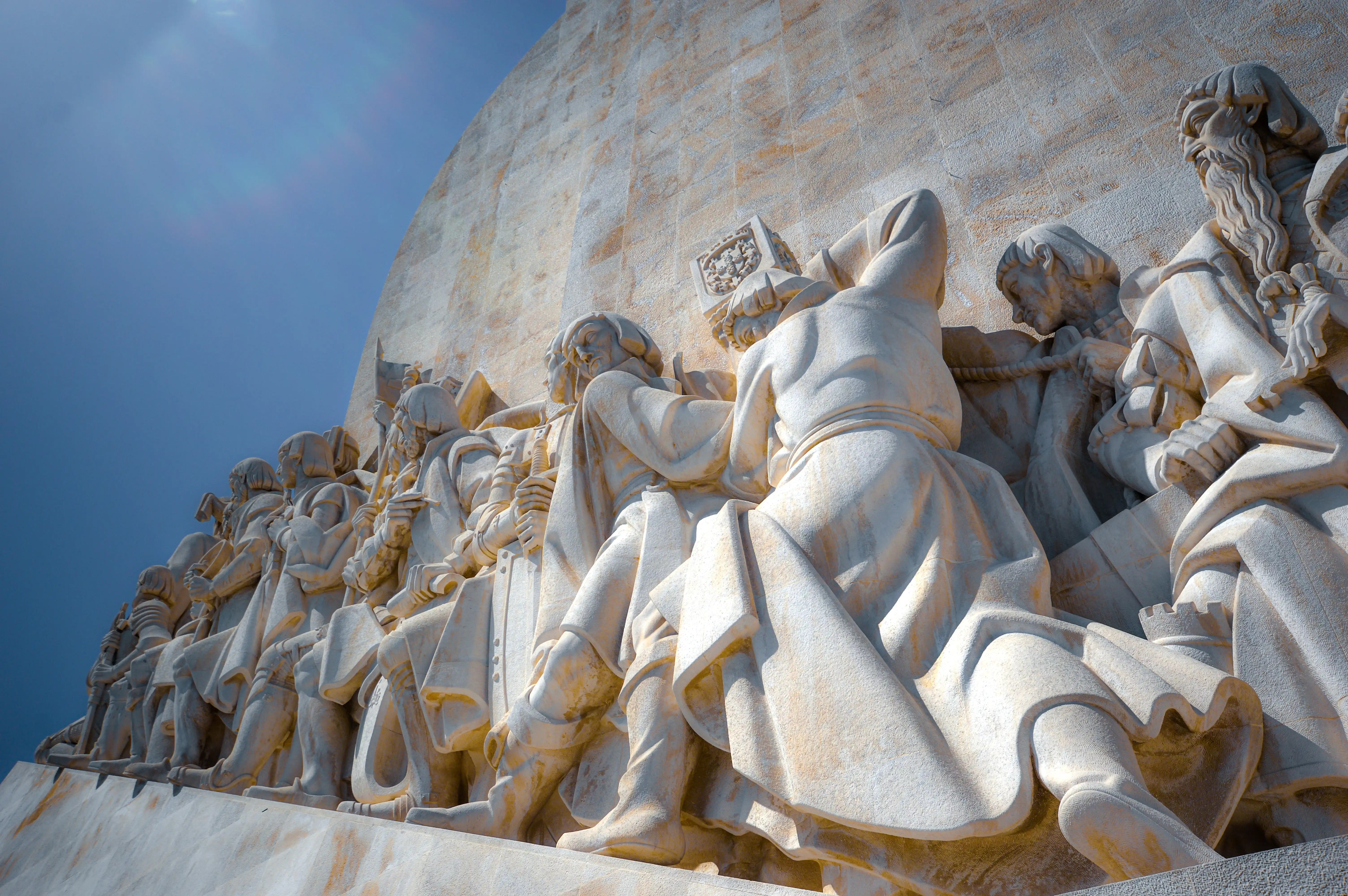 Close-up of a pale stone monument featuring a line of carved figures in historical dress, viewed from below against a bright blue sky with a sun flare.